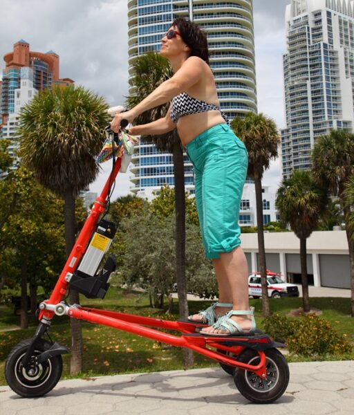 A woman wearing a bikini top and turquoise capri pants rides a stand-up red electric scooter in Palm Beach Gardens, Florida, with palm trees and tall modern buildings in the background.