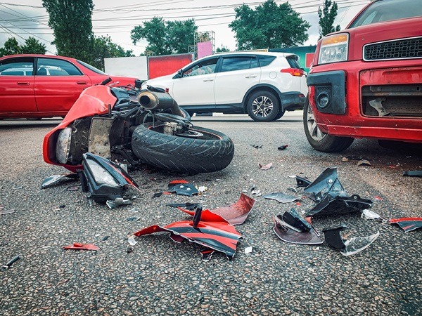 Red motorcycle lying on its side in the street after a crash, surrounded by broken debris and plastic fragments, with multiple cars stopped nearby.