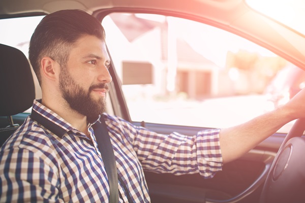 A man with a beard and a plaid shirt sitting in the driver's seat of a car, looking forward with his hands on the steering wheel.