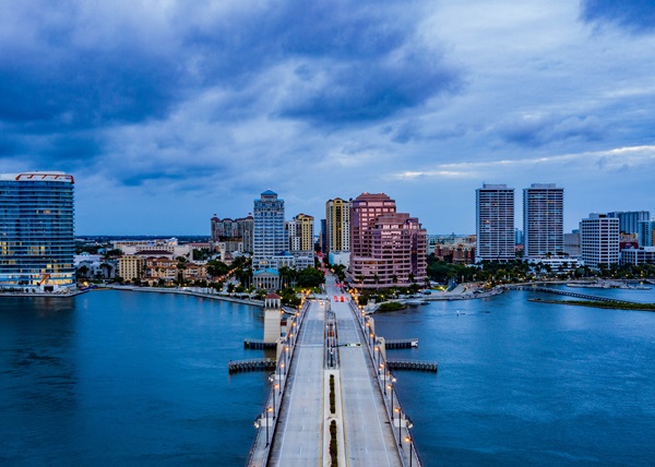 Aerial view of a bridge leading into downtown West Palm Beach, Florida with high-rise buildings and waterfront on both sides.